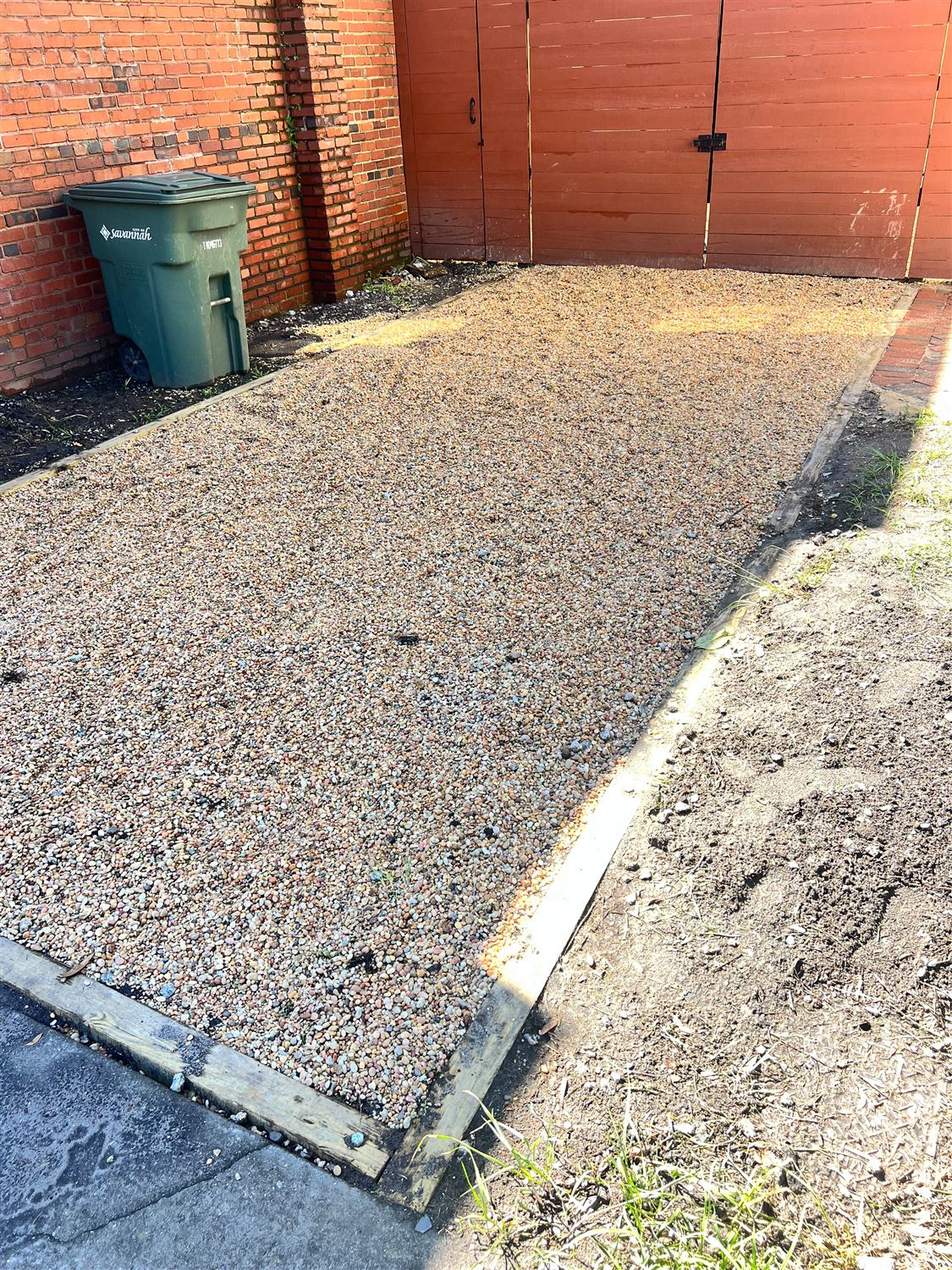 Gravel parking pad edged with pressure-treated timbers along a brick wall